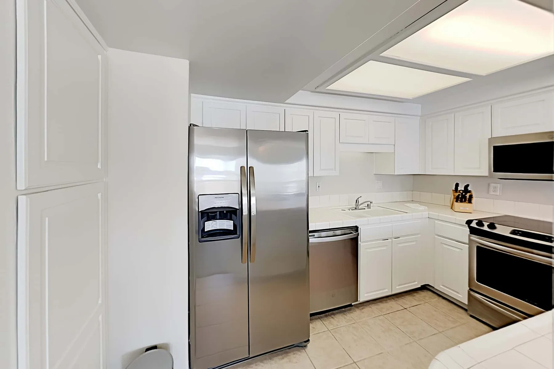 Una cocina luminosa con gabinetes blancos, electrodomésticos de acero inoxidable y piso de baldosas.