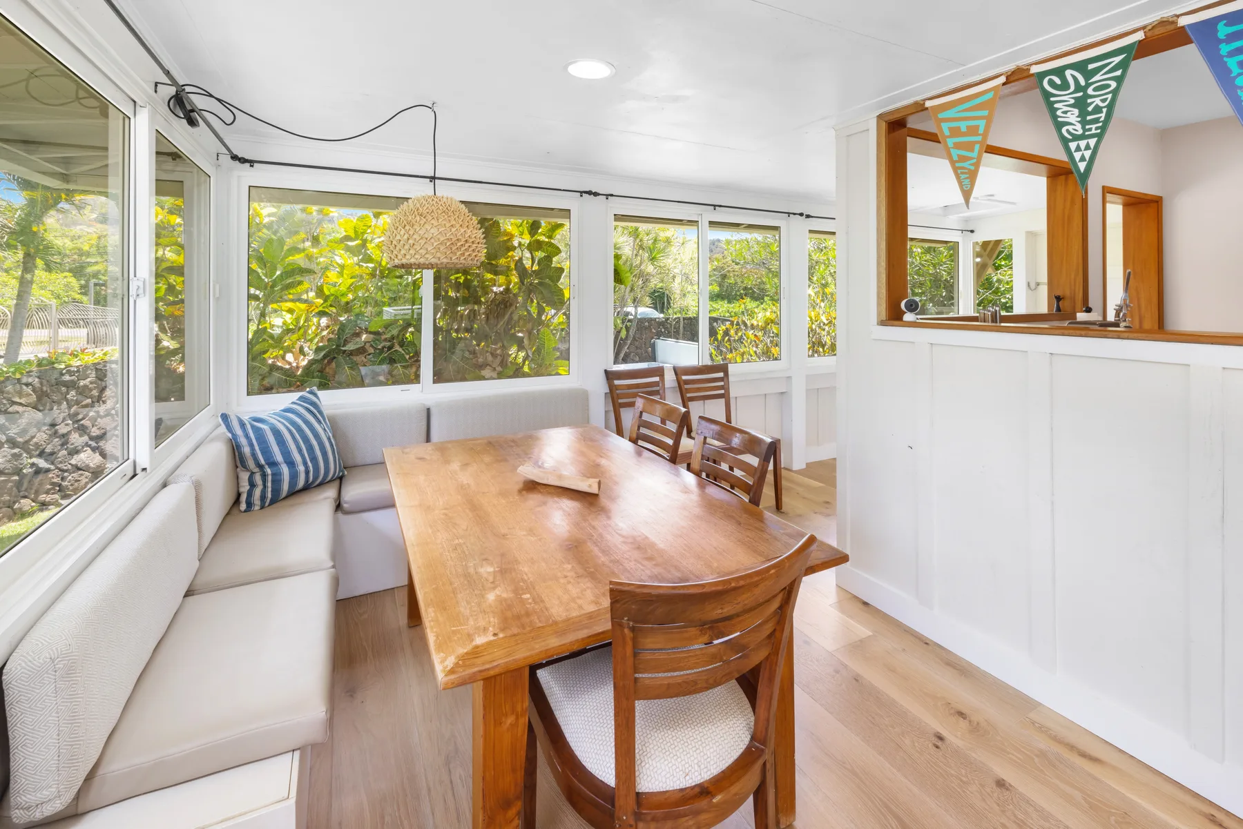 A bright dining area has a long wooden table beside windows overlooking greenery.