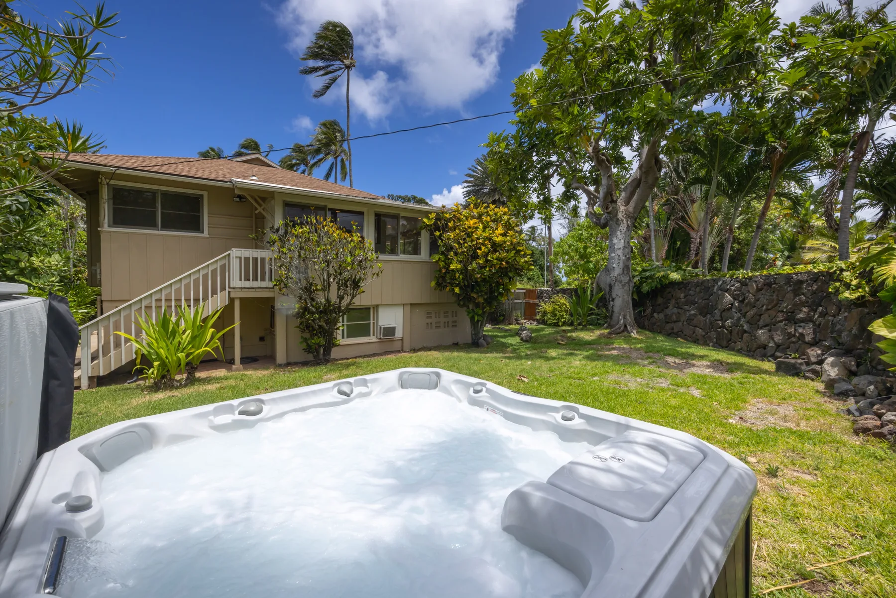 A backyard hot tub sits on a lawn beside a raised house and leafy trees.