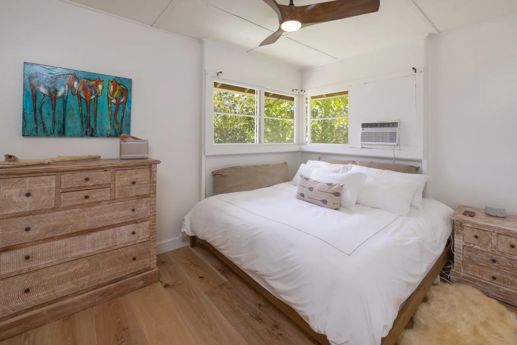 A bright bedroom with a bed beneath corner windows and wooden dressers.