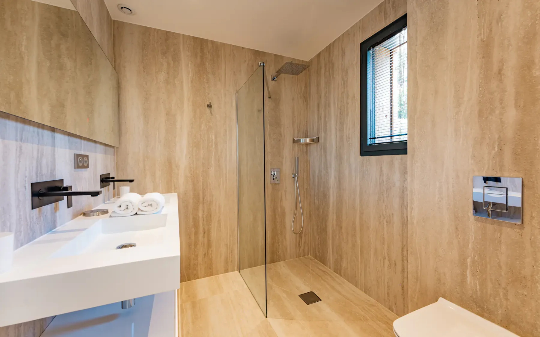 Baño con azulejos, ducha de vidrio, lavabo suspendido y una ventana con persianas.
