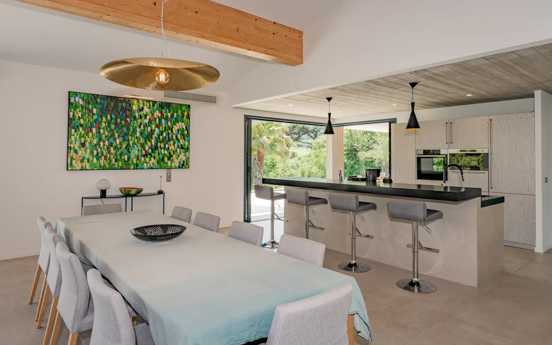 Cocina y comedor modernos de planta abierta con mobiliario de madera clara, una mesa larga, taburetes de barra, una ventana grande y obras de arte coloridas en la pared.