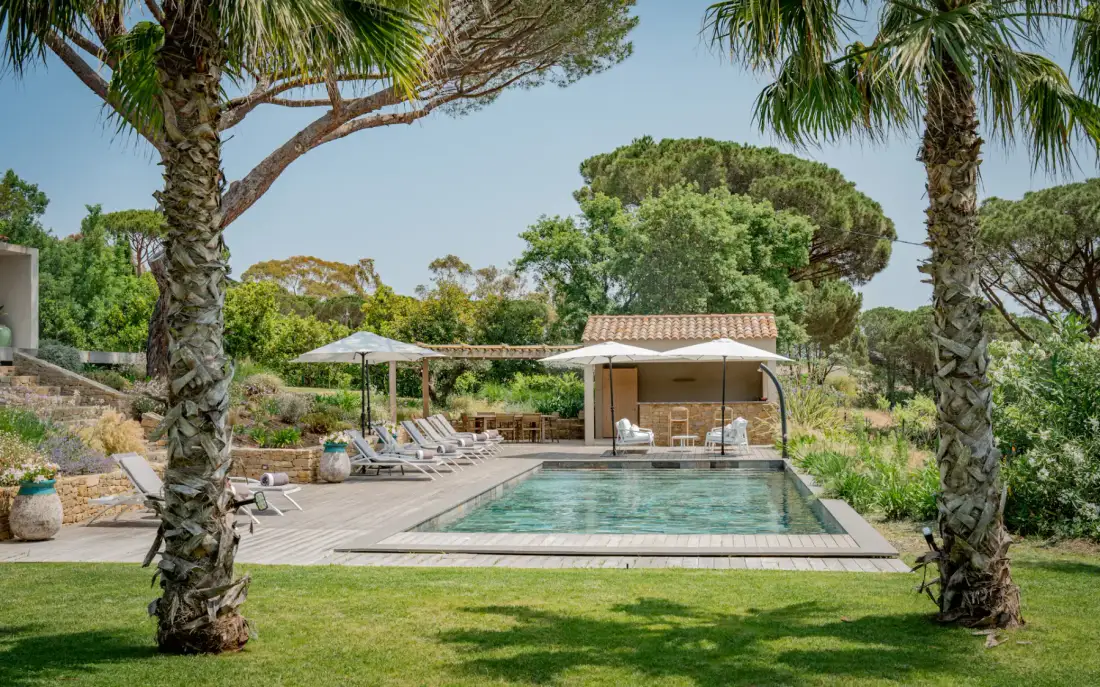Una tranquila zona de piscina al aire libre con tumbonas, sombrillas y un pequeño edificio, rodeada de exuberante vegetación y palmeras.