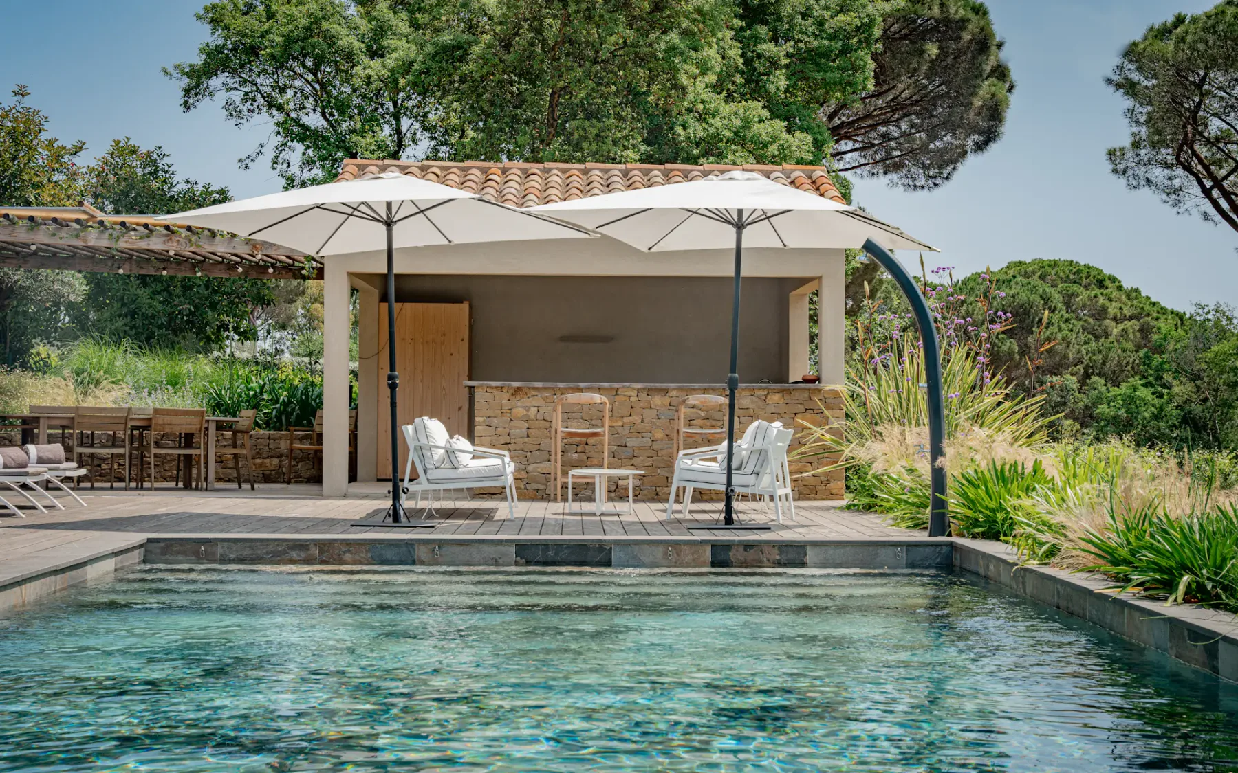 Una piscina al aire libre está junto a un patio sombreado con asientos y vegetación alrededor.