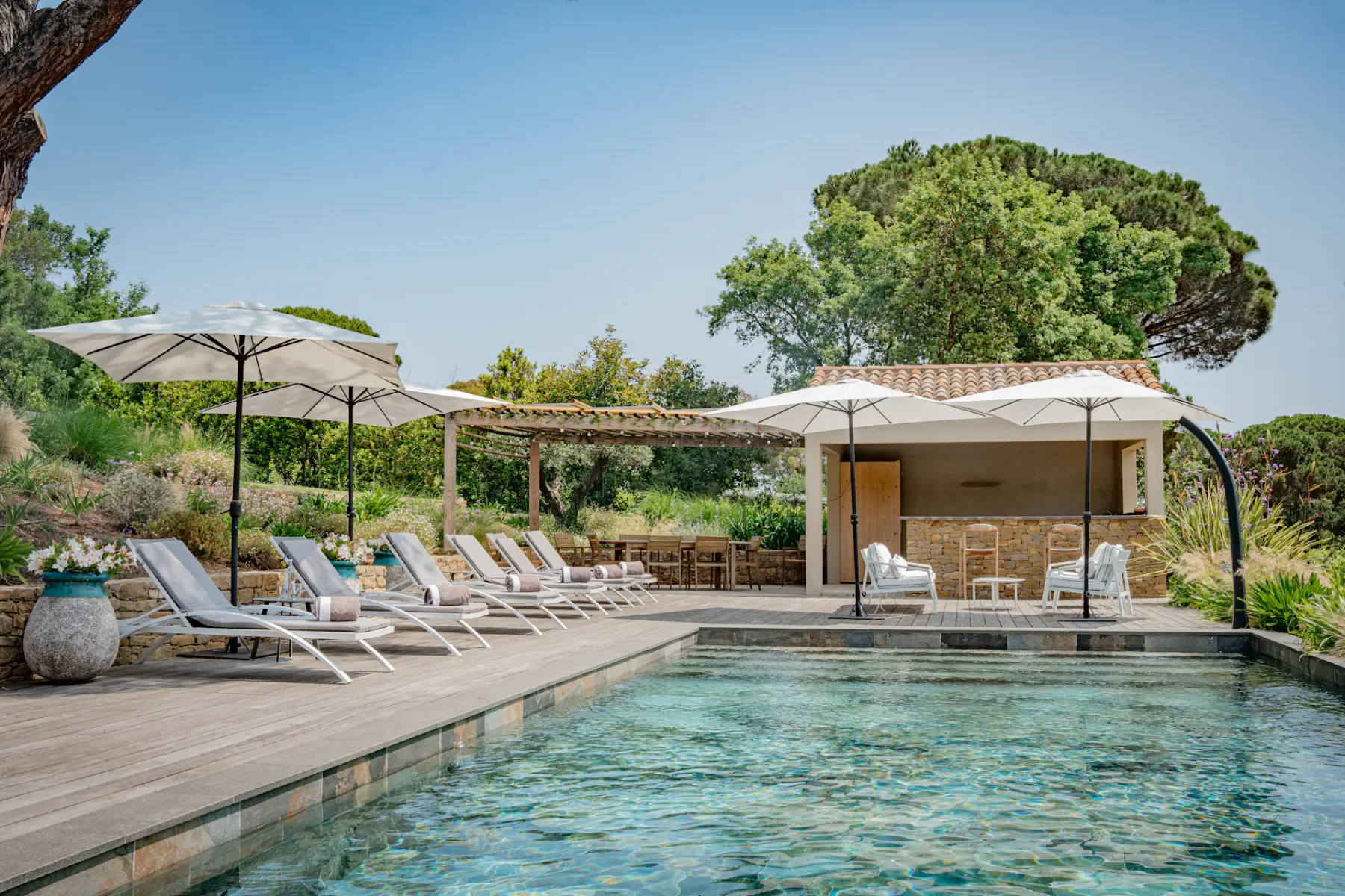 Una tranquila zona de piscina al aire libre con tumbonas, sombrillas y una cabaña cercana, rodeada de exuberante vegetación y bajo un cielo azul despejado.