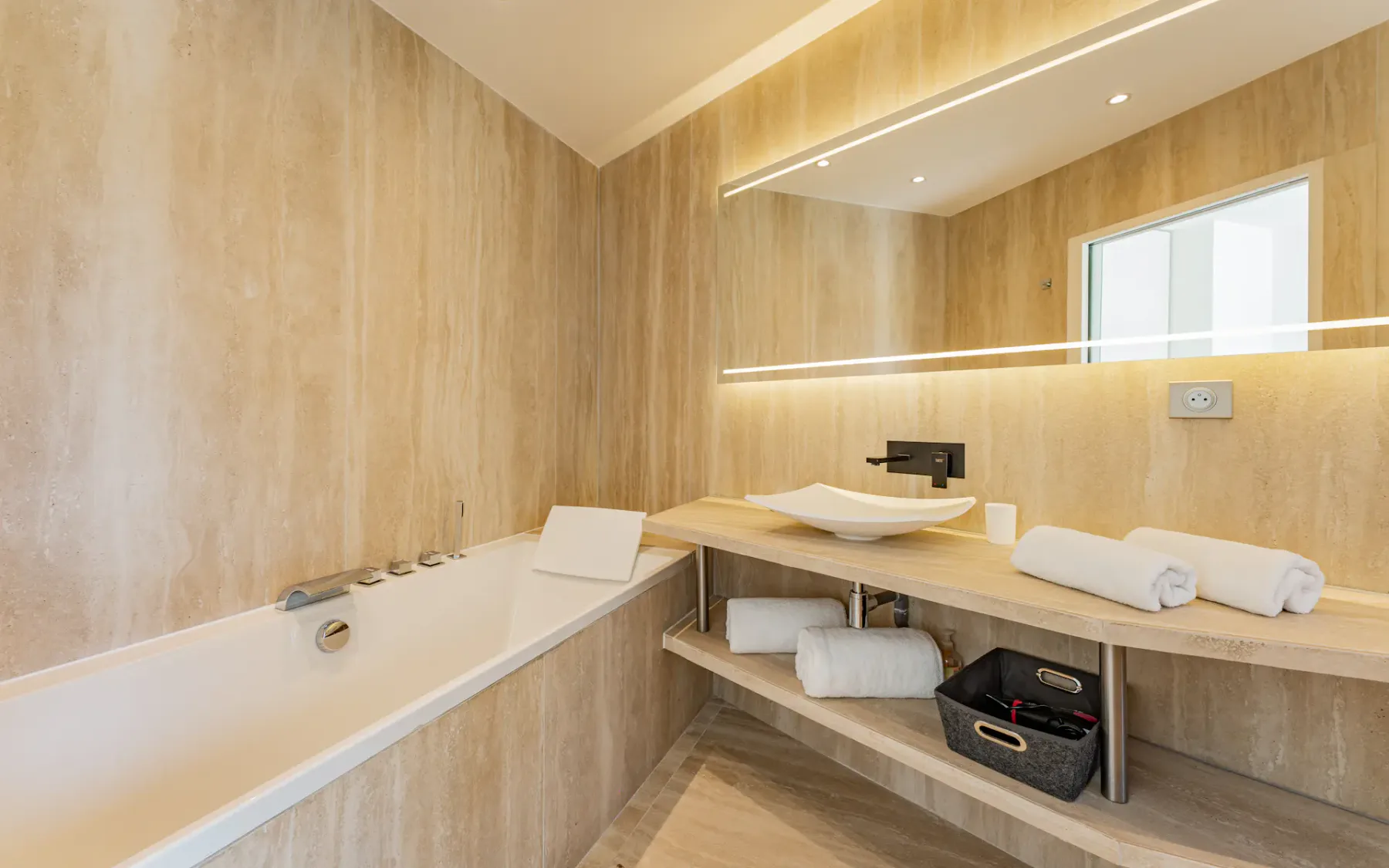 Baño moderno con bañera rectangular, un mueble de lavabo de madera con lavabo sobre encimera, toallas enrolladas, un espejo grande y decoración en tonos beige cálidos.