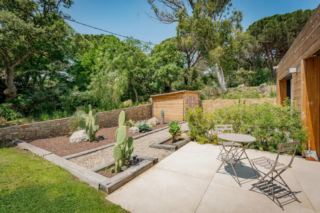 Un patio de jardín soleado con una mesa pequeña y sillas, cactus en un bancal elevado, un cobertizo de madera y árboles frondosos al fondo.