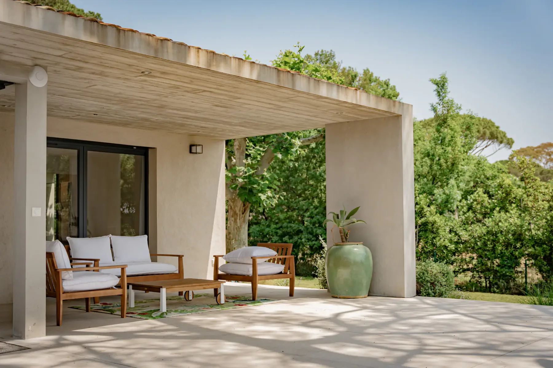 Patio exterior con sillas de madera con cojines blancos, una mesa pequeña y una gran maceta verde. Una cubierta proporciona sombra. Exuberante vegetación al fondo.
