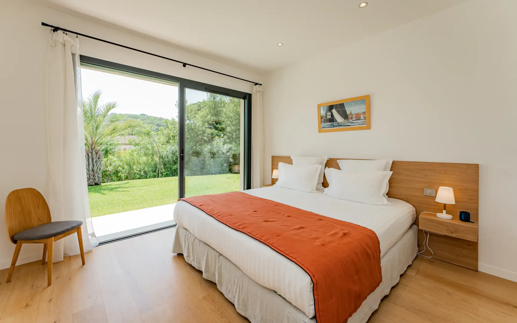 Dormitorio luminoso con una cama grande, una manta decorativa naranja, suelo de madera, una pequeña lámpara de noche y vistas a la vegetación a través de una puerta corredera de cristal.