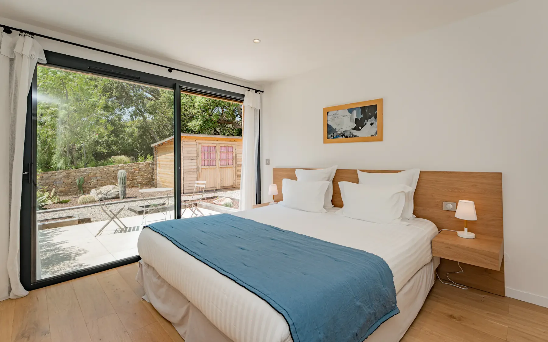 Dormitorio moderno con una cama grande, una manta azul, cabecero de madera, lámparas pequeñas y vistas a un jardín a través de puertas correderas de cristal.