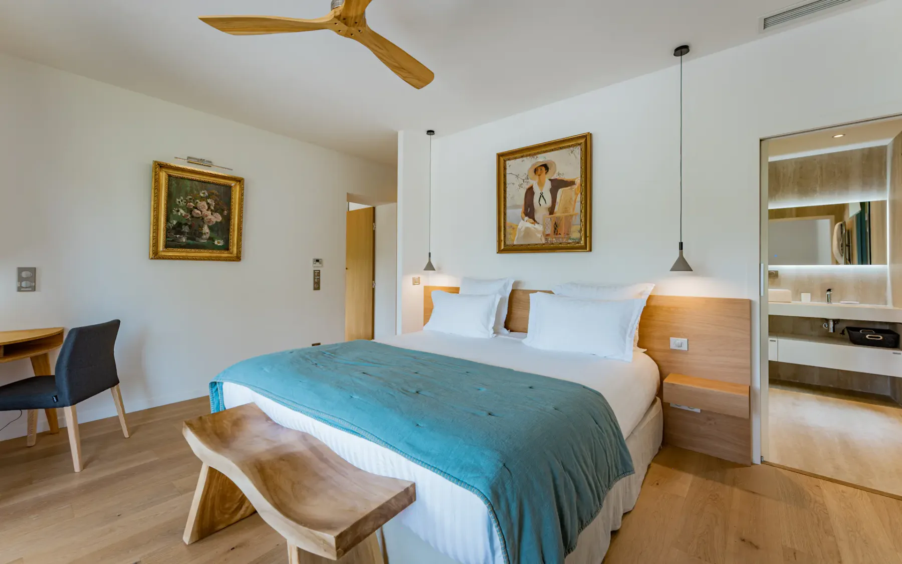Habitación de hotel moderna con cama doble, manta verde azulada, banco de madera, escritorio, arte en las paredes, lámparas colgantes y vistas abiertas al baño.