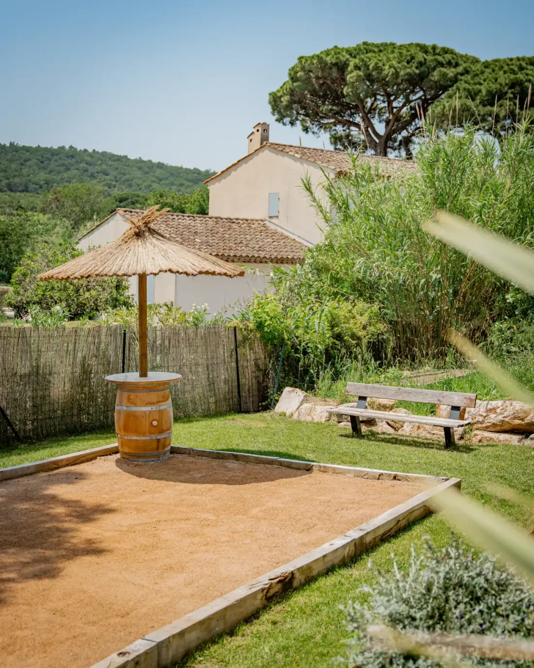 Un jardín soleado con una pista de bochas de arena, una sombrilla de paja y un banco de picnic.