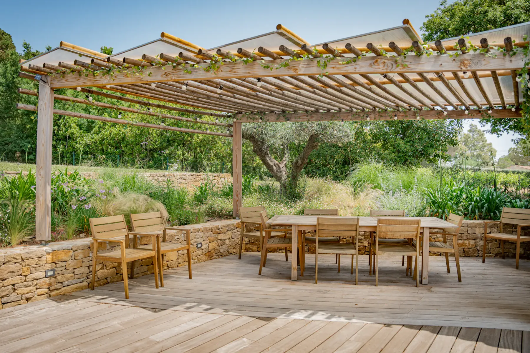 Patio exterior con sillas y mesas de madera bajo una pérgola, rodeado de vegetación y muros de piedra.