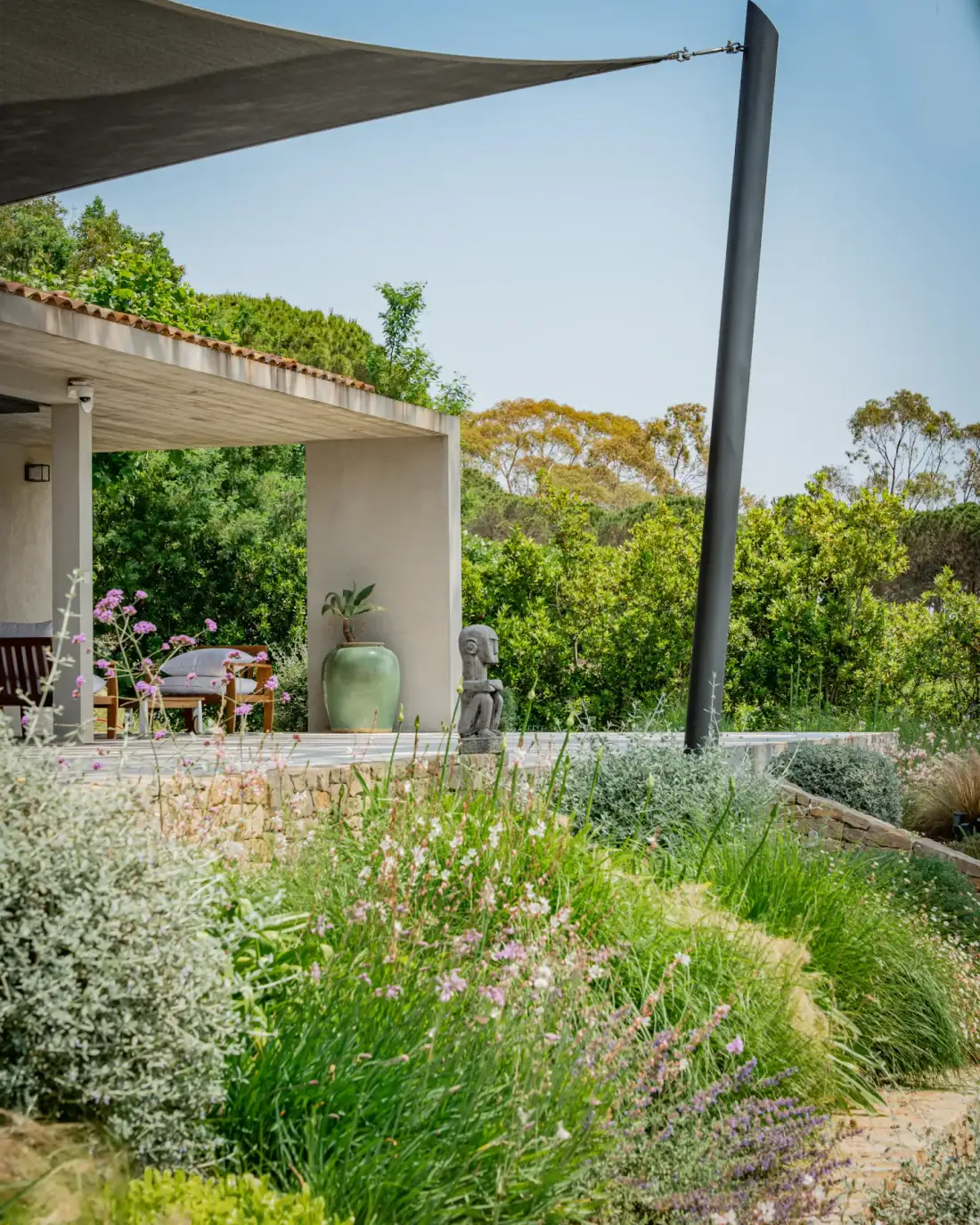 Un jardín frondoso junto a un patio sombreado con asientos al aire libre bajo una cubierta de tela.