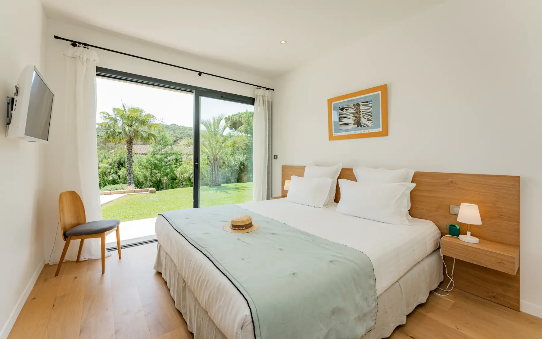 Dormitorio luminoso con una cama grande, suelo de madera clara y una puerta corredera de cristal que da a un frondoso jardín. Televisor montado en la pared y una lámpara pequeña.