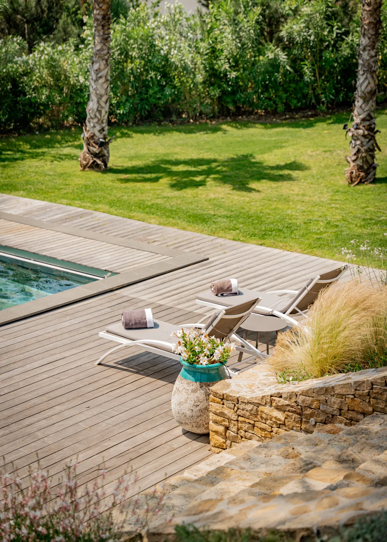 Una terraza junto a la piscina con tumbonas, césped y árboles.