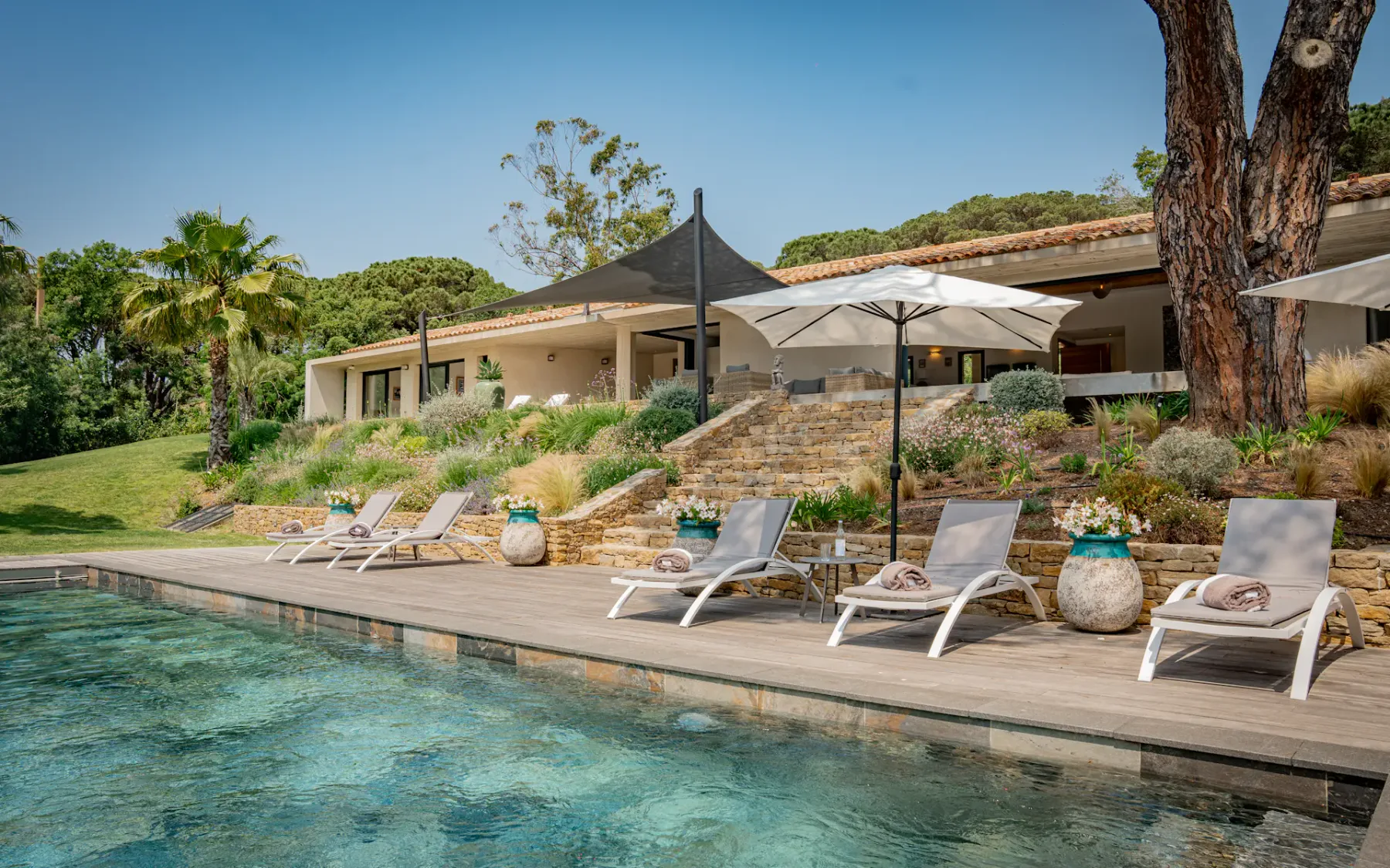 Un patio junto a la piscina con tumbonas y toldos, junto a una casa moderna.