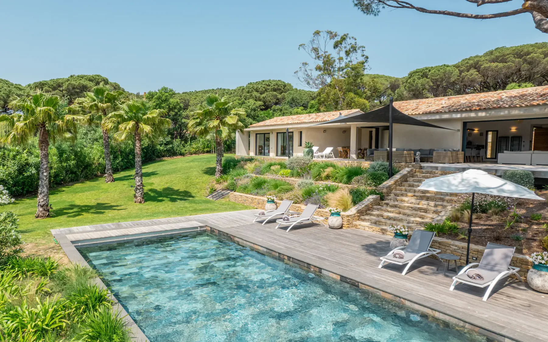 Una villa moderna con piscina, rodeada de palmeras y exuberante vegetación, bajo un cielo azul despejado.