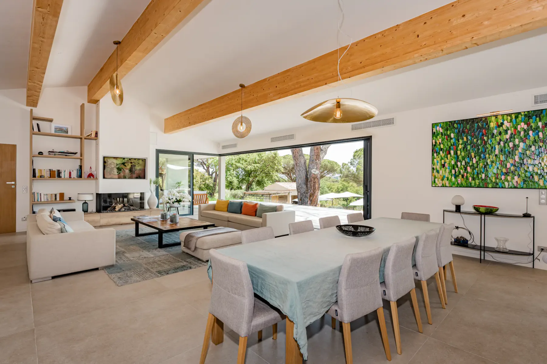 Amplia zona de salón y comedor de planta abierta con grandes ventanas, vigas de madera, mobiliario moderno y obras de arte coloridas en la pared.