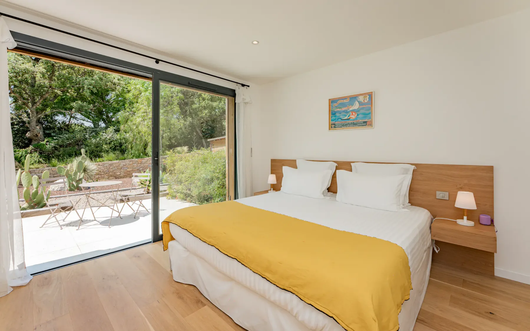 Dormitorio luminoso con una cama grande, una manta amarilla, lámparas de noche, un cuadro y puertas de cristal que dan a un patio con vegetación en el exterior.