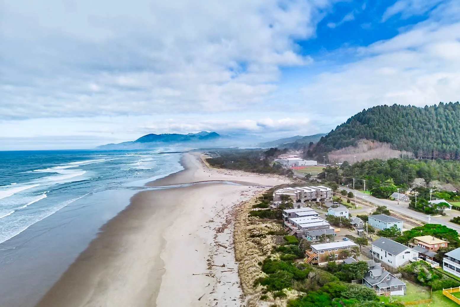 Vista aérea de la costa con una playa de arena, olas y casas junto a una colina boscosa.