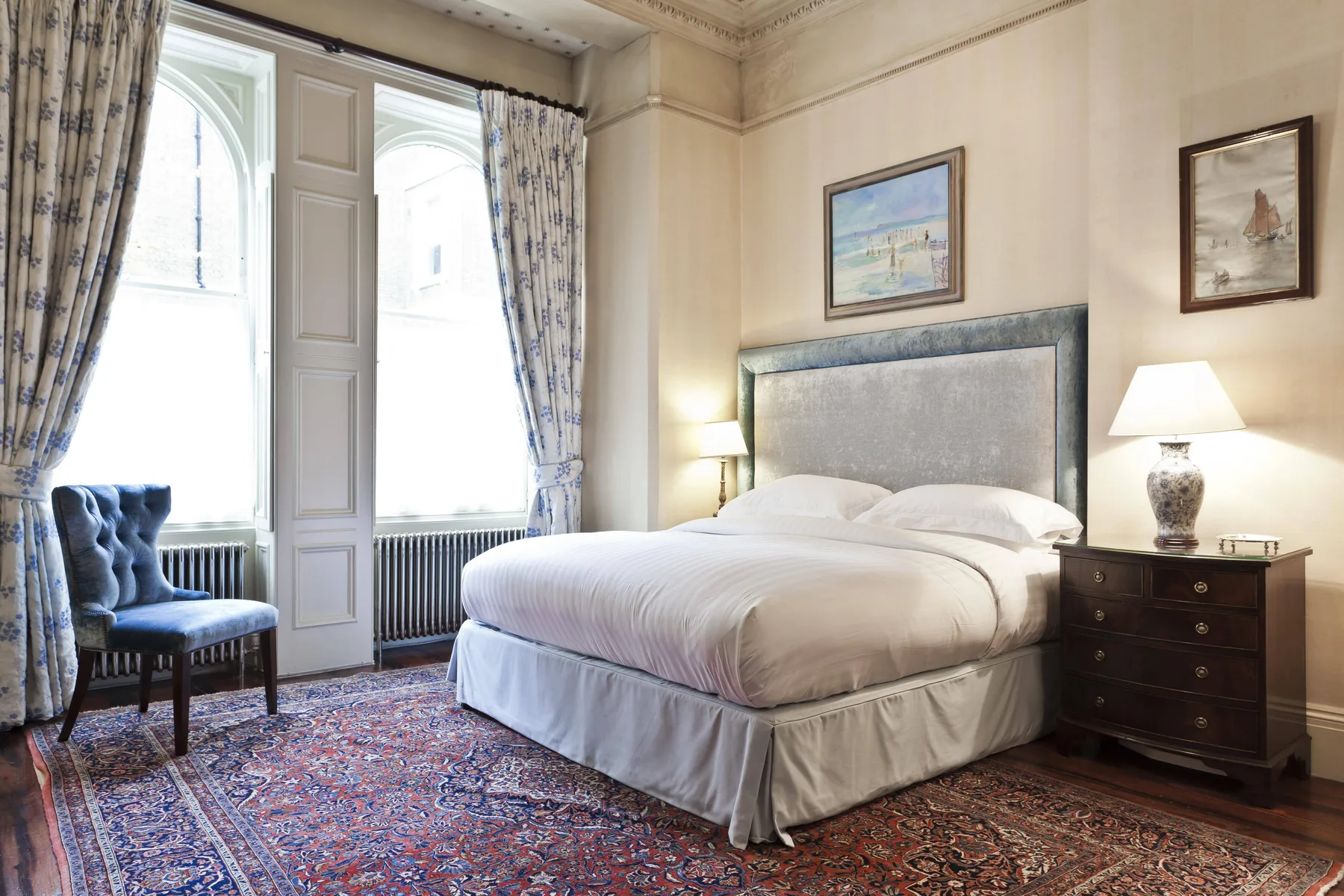 Un dormitorio con una cama grande, ventanas altas con cortinas y una alfombra estampada.