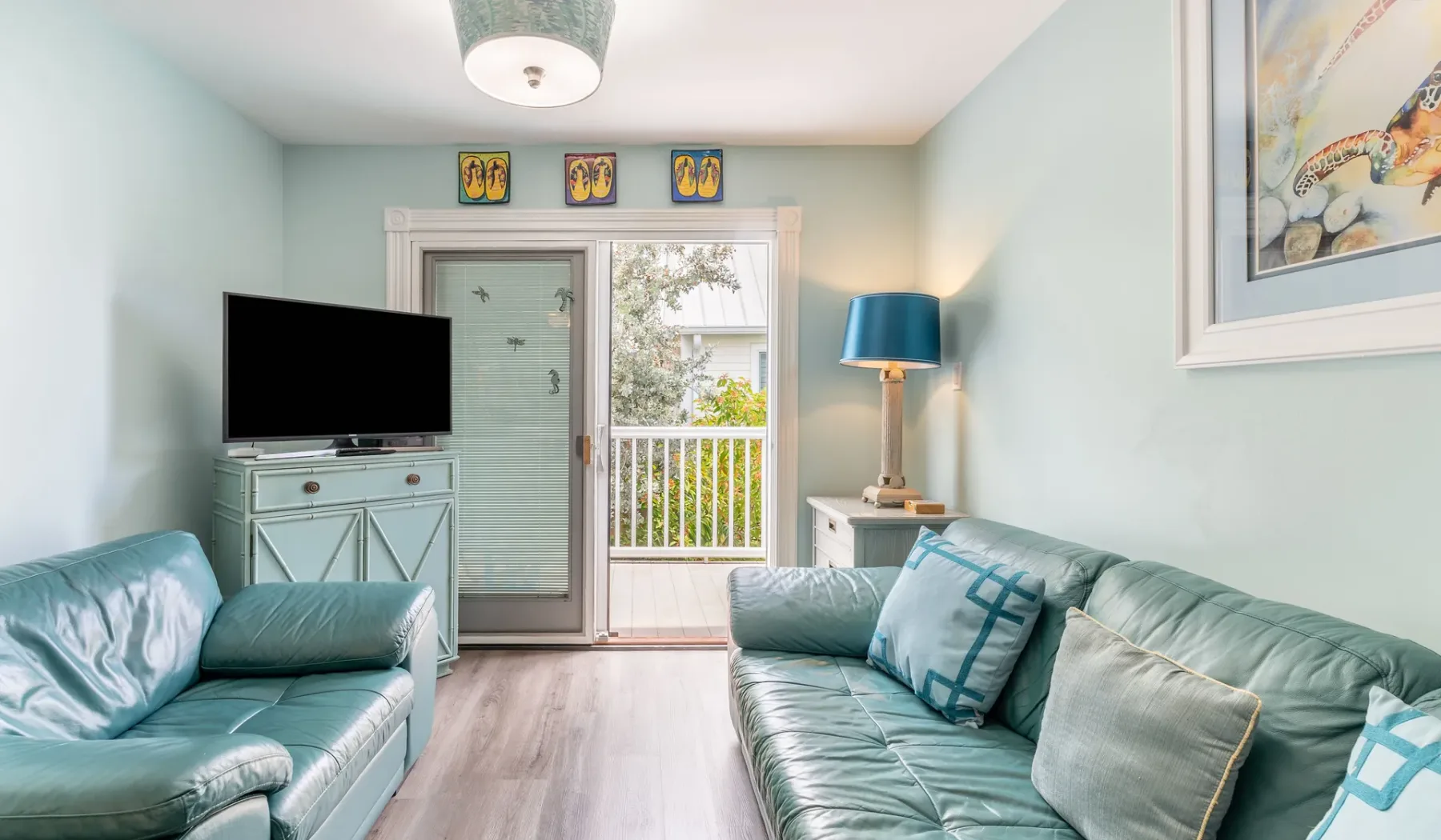 Una sala luminosa con asientos color turquesa, un televisor y una puerta de vidrio que da a un balcón.