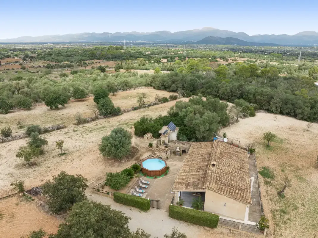 Vista aérea de una casa rural rodeada de árboles, con montañas al fondo.