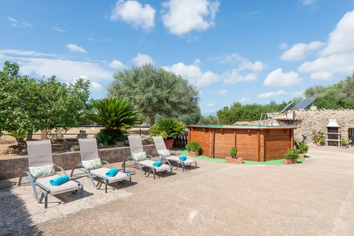 Un patio exterior tiene asientos reclinables junto a una bañera de hidromasaje de madera y un jardín.