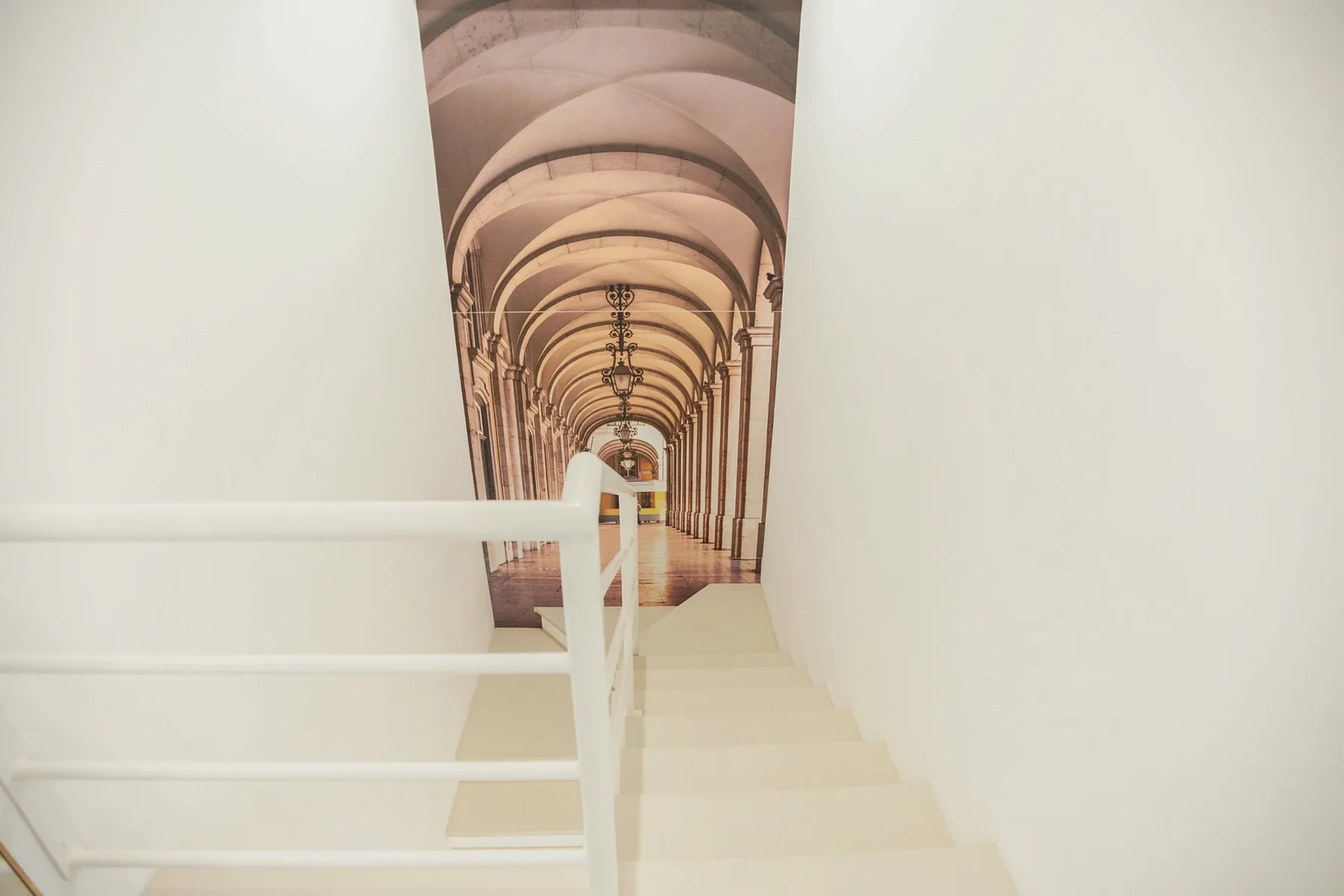 A white staircase with a simple railing stands beside a tall arched corridor mural.