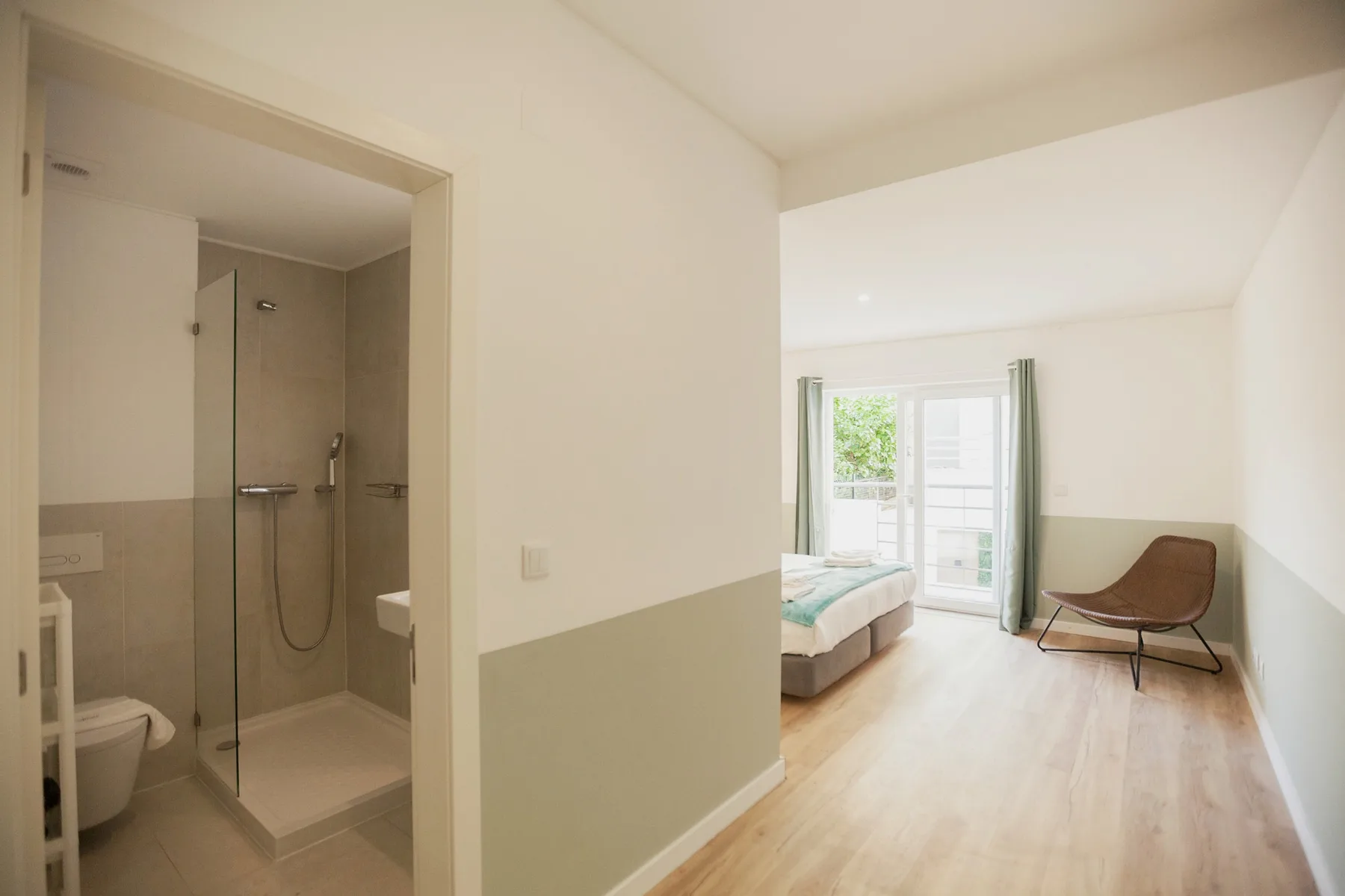 A bedroom with a bed and chair beside a bathroom with a shower.