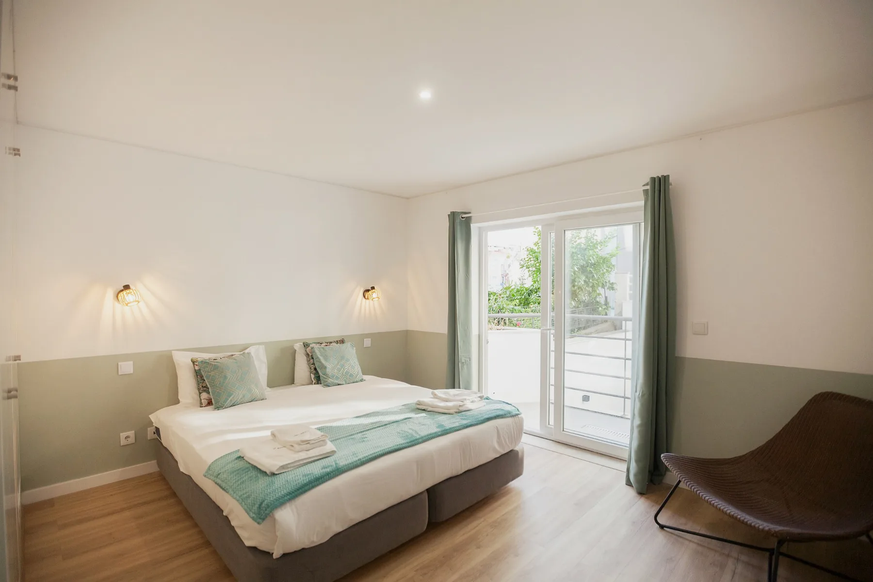 A bright bedroom with a double bed, bedside lights, and a glass door opening to a balcony.