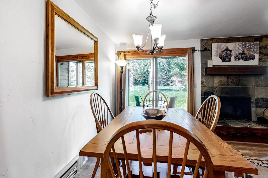 A wooden dining table and chairs sit beside large windows overlooking greenery.