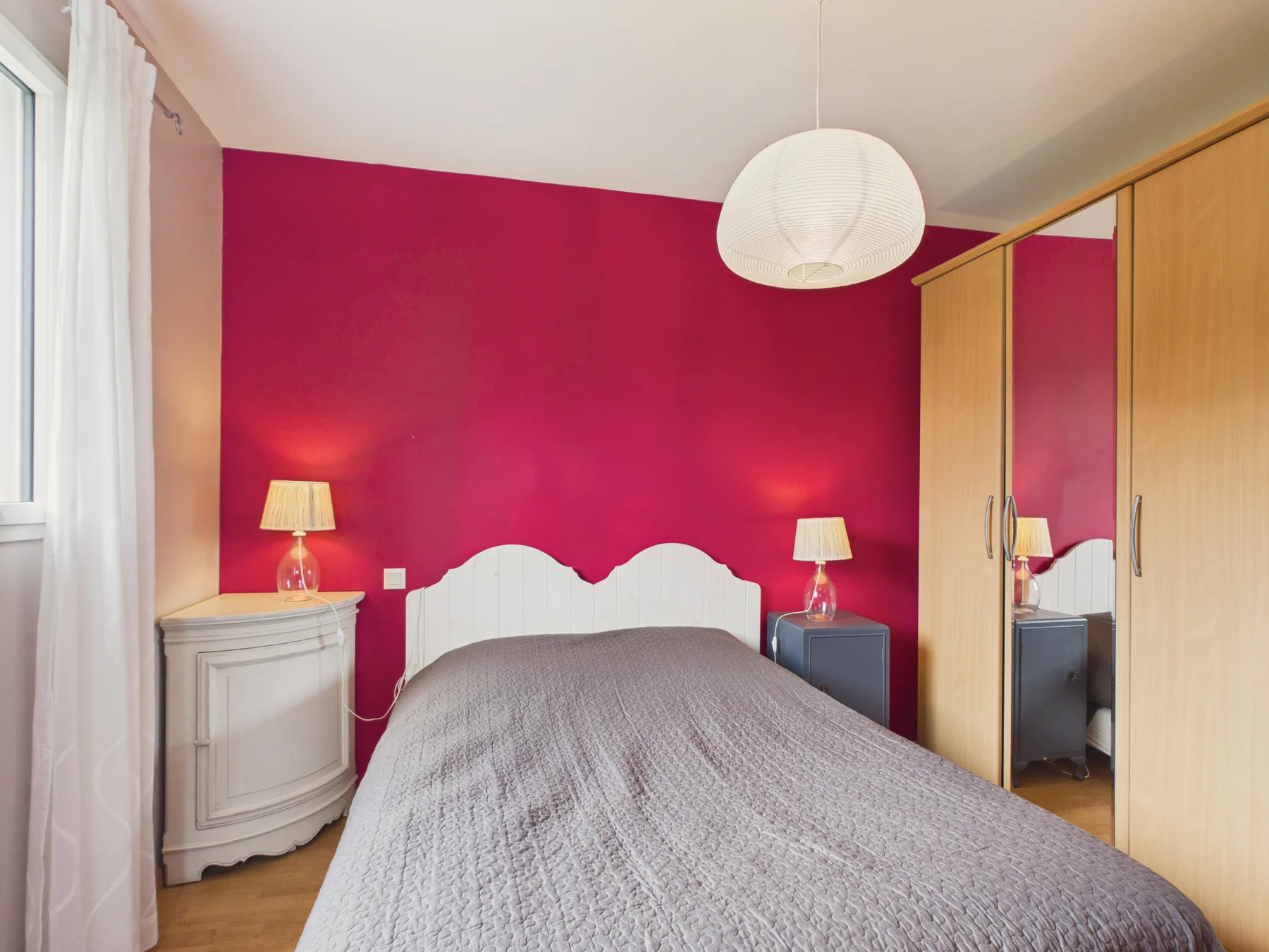 A bedroom with a bed against a bright pink wall and a mirrored wardrobe.