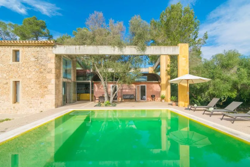 Una piscina frente a una casa moderna con fachada de vidrio y tumbonas cerca.