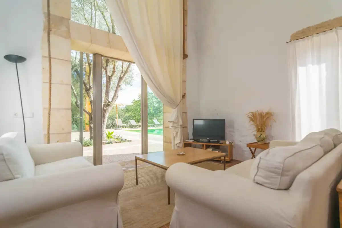 Una sala de estar luminosa con asientos claros y grandes ventanales hacia un jardín.