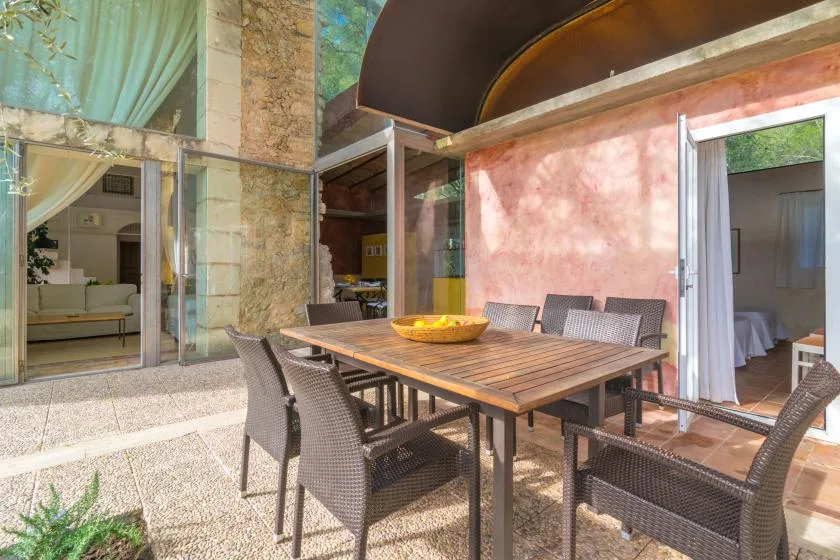 Un comedor en el patio con una mesa de madera y asientos trenzados junto a puertas de vidrio abiertas.