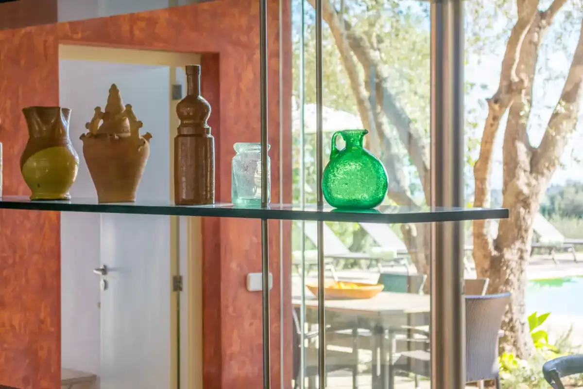 Una estantería de vidrio con cerámica decorativa está junto a una ventana luminosa.