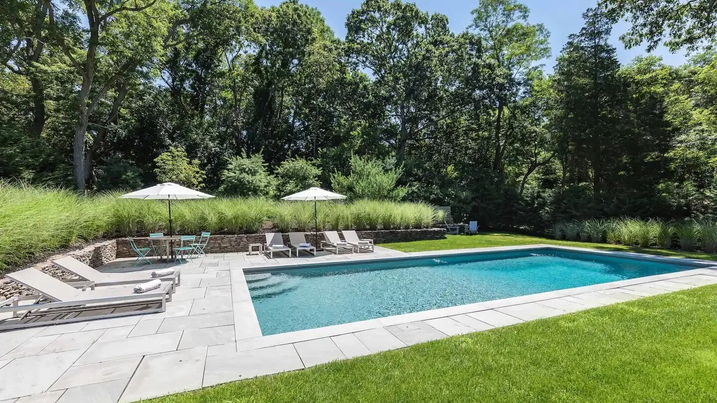 Una piscina está rodeada por una terraza pavimentada, asientos y césped verde.
