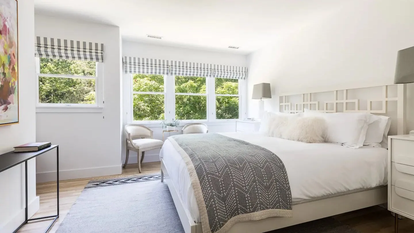 Un dormitorio luminoso con ropa de cama blanca, una manta estampada y ventanas con vistas a la vegetación.