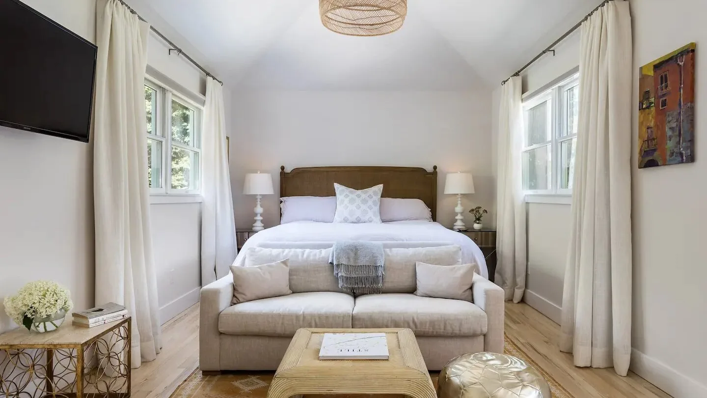 Un dormitorio luminoso con una cama, asientos y ventanas altas con cortinas.