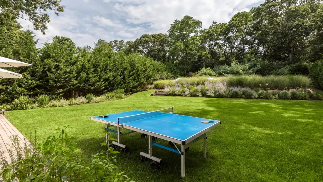 Una mesa azul de ping-pong está sobre un césped rodeado de árboles y plantas con flores.