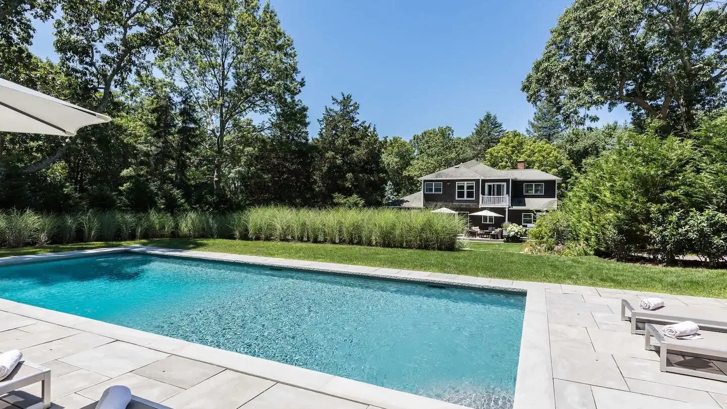 Una piscina al aire libre está junto a un patio pavimentado, con una casa y árboles al fondo.