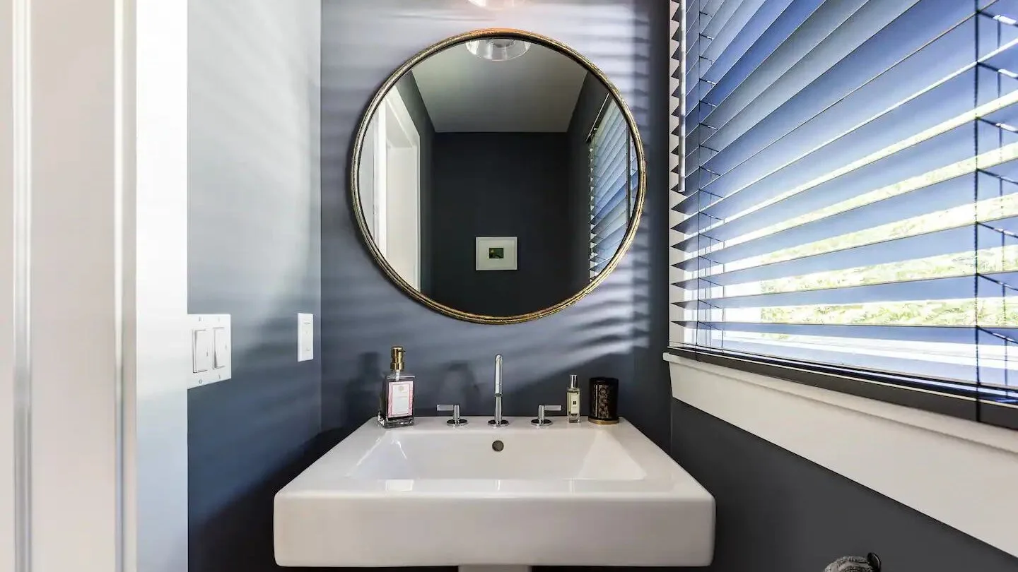 Un lavabo de baño con un espejo redondo y persianas en la ventana.
