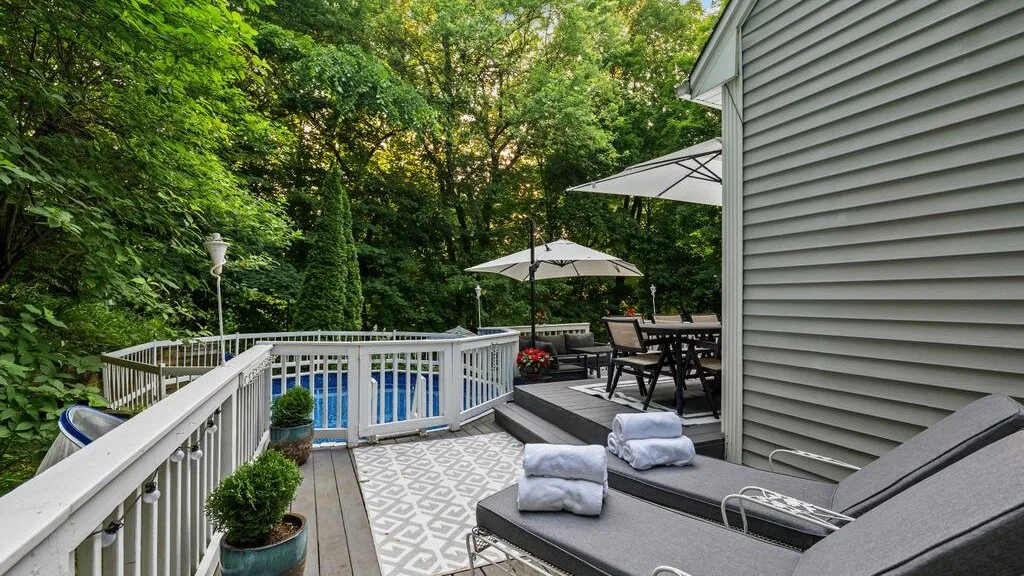 An outdoor deck with seating and umbrellas overlooks a swimming pool surrounded by trees.
