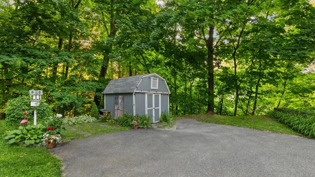 A small gray shed stands beside a paved driveway in a leafy yard.