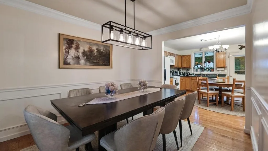 A dining area with a long table and upholstered seating opens onto a kitchen.
