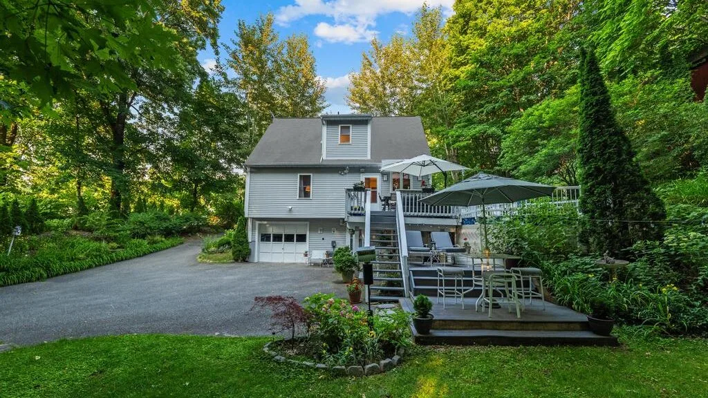 A house and raised outdoor deck sit among trees beside a paved driveway.
