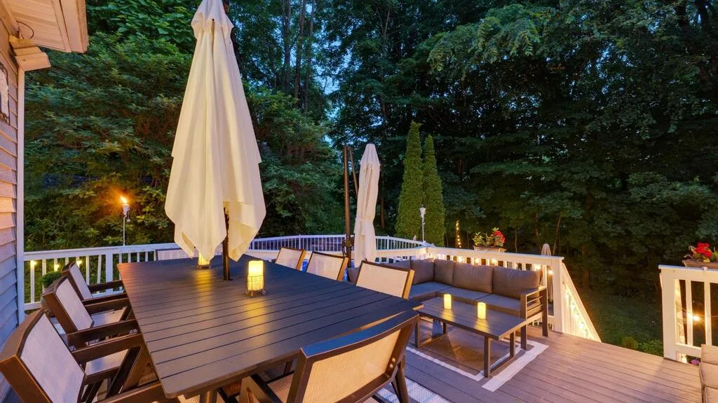 An outdoor deck has a dining table, seating, and closed umbrellas among trees.