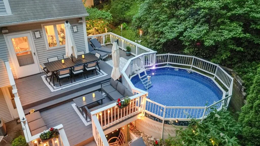 A raised outdoor deck with seating overlooks a fenced swimming pool surrounded by greenery.